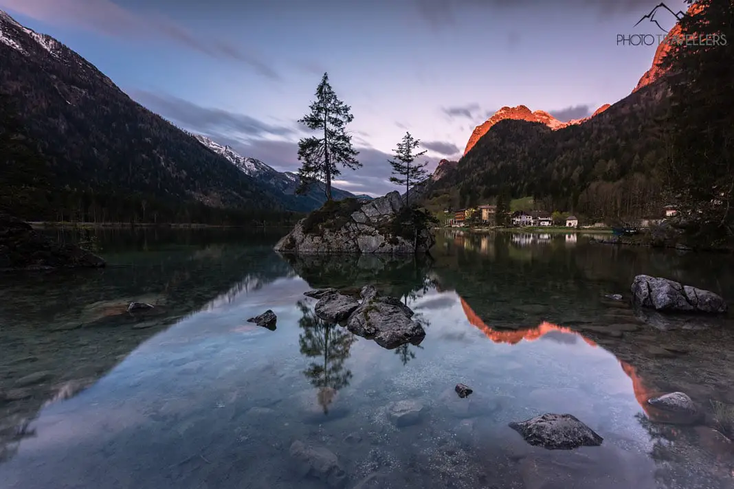 Der bekannte Fels mit Bäumen im Hintersee im Berchtesgadener Land. Am Morgen werden die Berge rot angestrahlt und spiegeln sich im Wasser