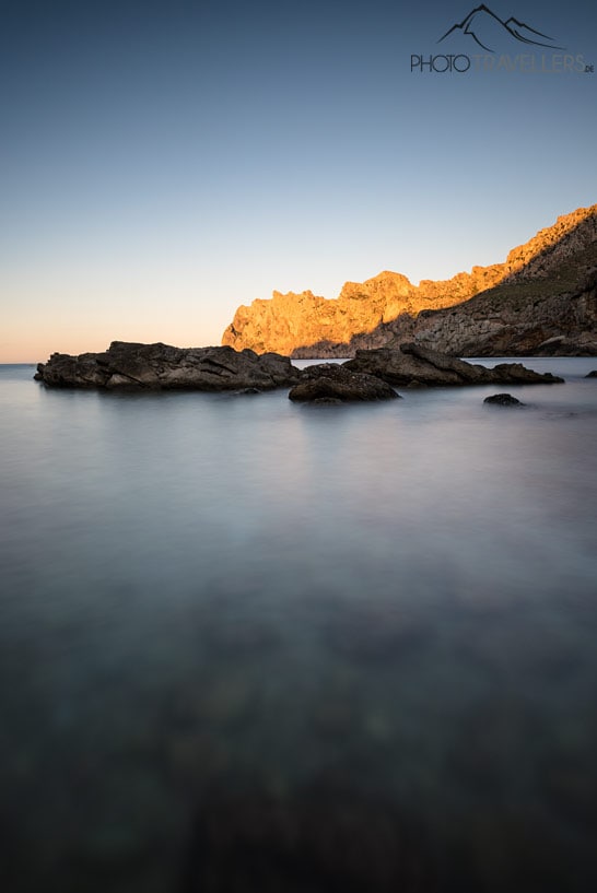 Der Strand von Cala Sant Vicenç mit Blick auf hohe Felswände, die am Abend von der Sonne angestrahlt werden