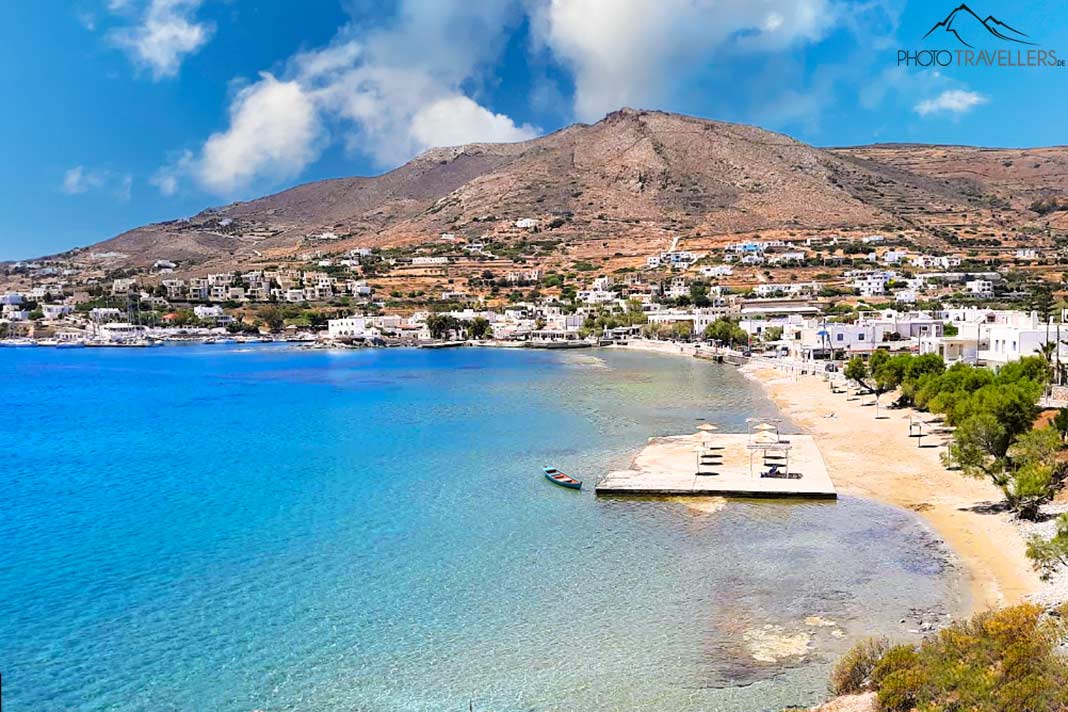 Schmaler Sandstreifen des Finikas Beach mit weißen Häusern und kahlem Berg im Hintergrund bei Sonnenschein