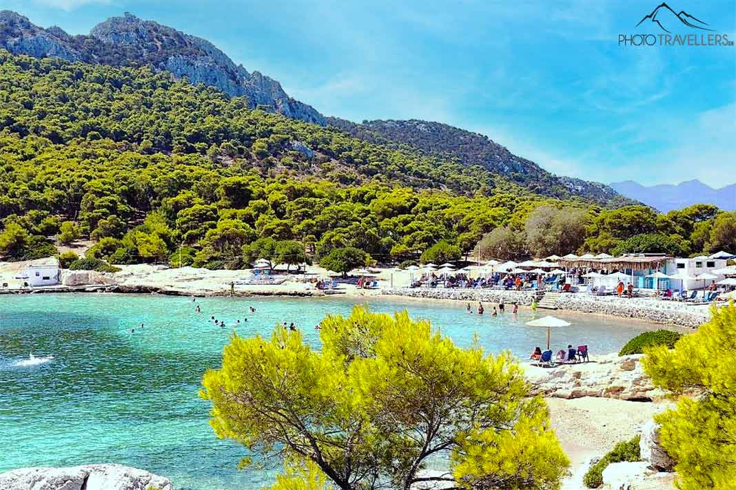 Badendes tummeln sich im türkisblauen Wasser vor den Felsen und bewaldeten Hügel der Insel Moni