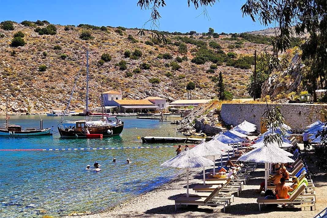 Sonnenliegen und Schirme reihen sich  vor Booten am Mandraki-Beach aneinander