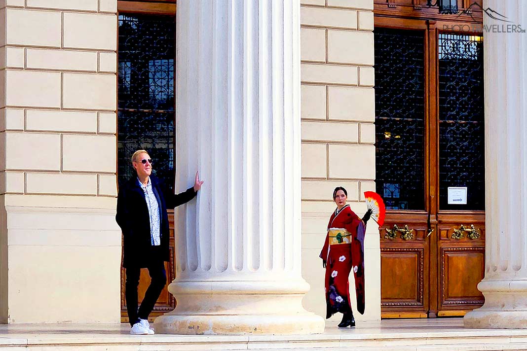 Ein Mann lehnt an einer Marmorsäule. Auf der anderen Seite steht eine Geisha in rotem Kostüm