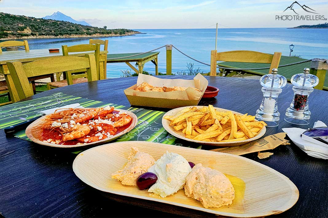 Mehrere Teller mit griechischen Speisen stehen auf einem Tisch mit Blick auf das Meer