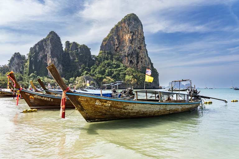 startseite-honeymoon-reiseziele Langboote an einem Strand in Thailand mit Felsen im Hintergrund