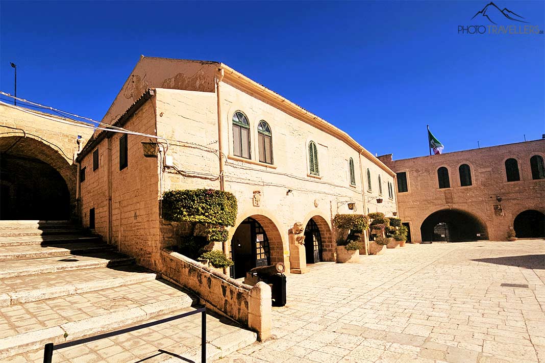 Mehrere Gebäude aus hellrotem Stein und ein gepflasterter Hof im Castello Aragonese