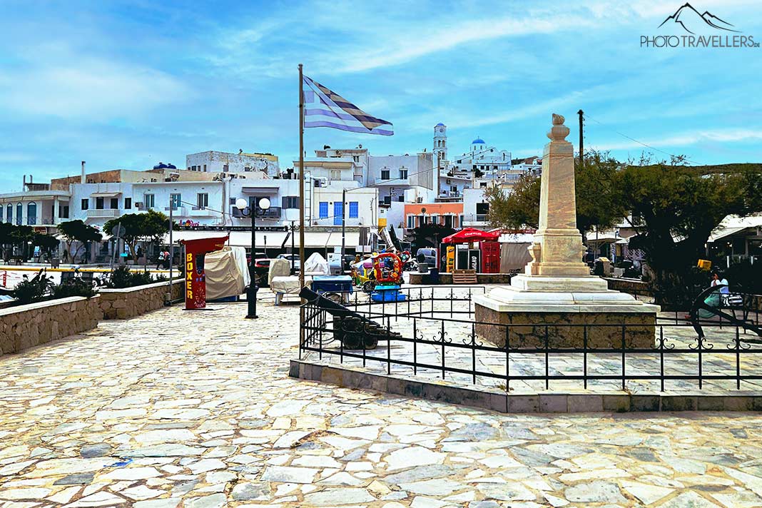 Die Promenade von Adamas mit Griechenland-Flagge und einem weißen Obelisken unter blauem Himmel
