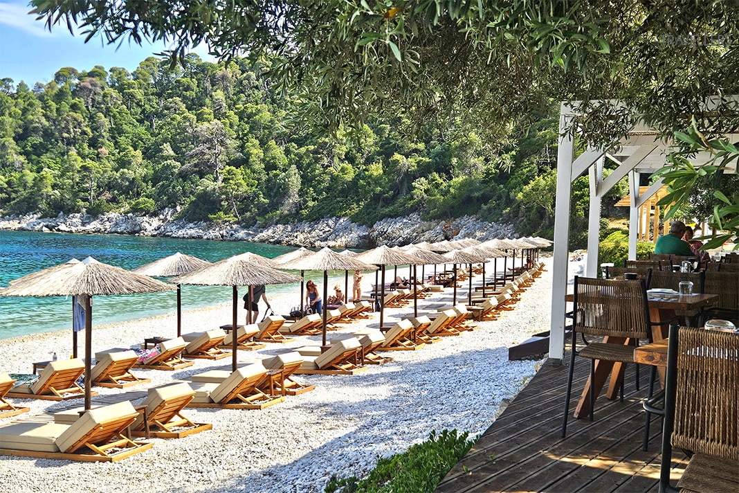 Aufgereihte Liegen  und Sonnenschirme am Strand Leftos Gialos mit Bäumen dahinter