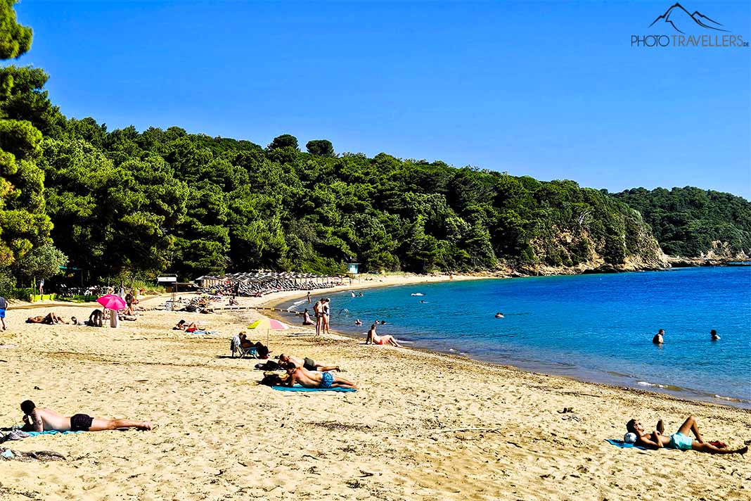 Mehrere Menschen liegen auf Handtüchern am Sandstrand von Agia Eleni, dahinter ist Wald