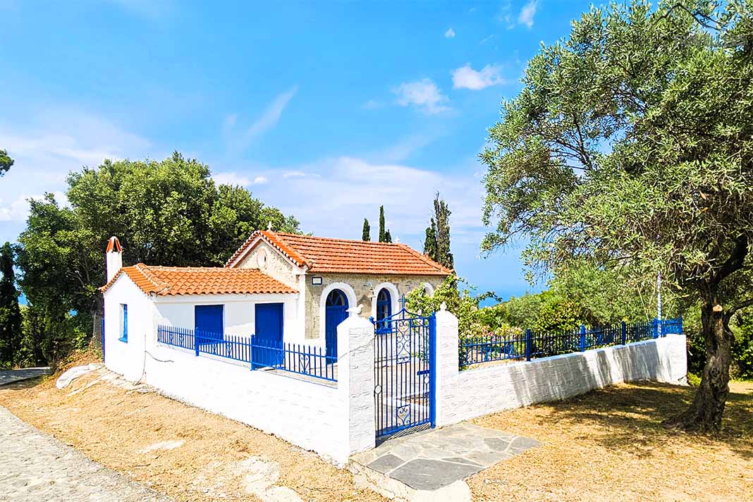 Steinkapelle Agios Theodoros mit roten Dachziegeln und blauen Türen zwischen Bäumen