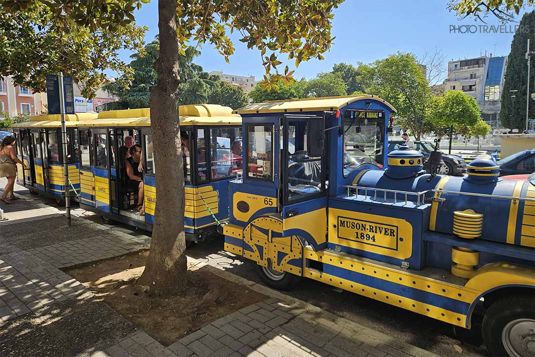 Die in gelb und blau lackierte Tram von Kavala mit Lok und Waggon vor einem Baum