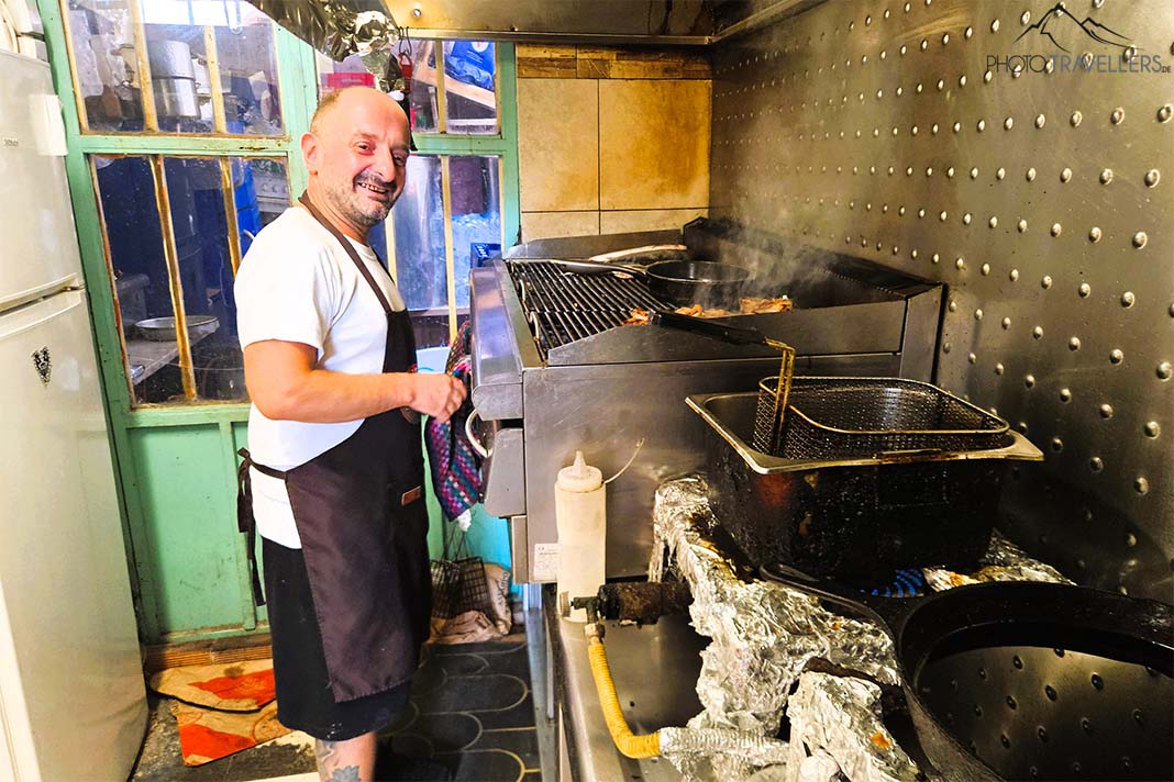 Ein Mann in Kochschürze steht vor einem Grillrost und lacht in die Kamera